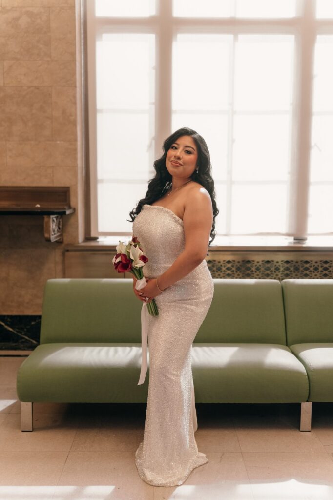 A bride holds a bouquet while wearing a sparkly white strapless gown while she awaits her NYC City Hall elopement ceremony. 