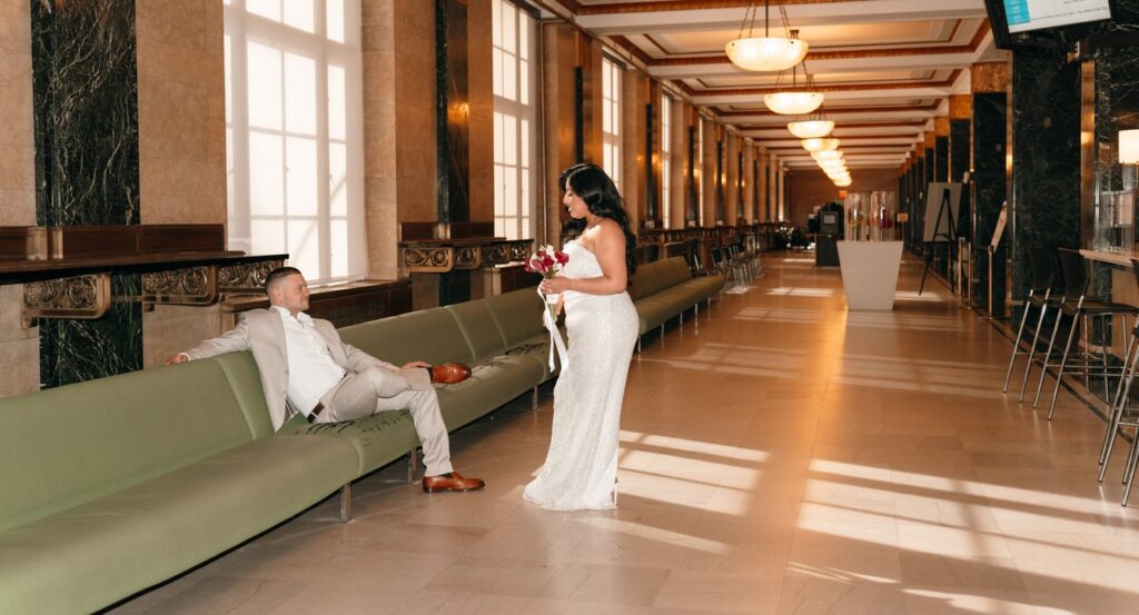 A bride stands before her groom, seated on a green couch in the waiting area of NYC City Hall in a photo taken by a NYC elopement photographer. 