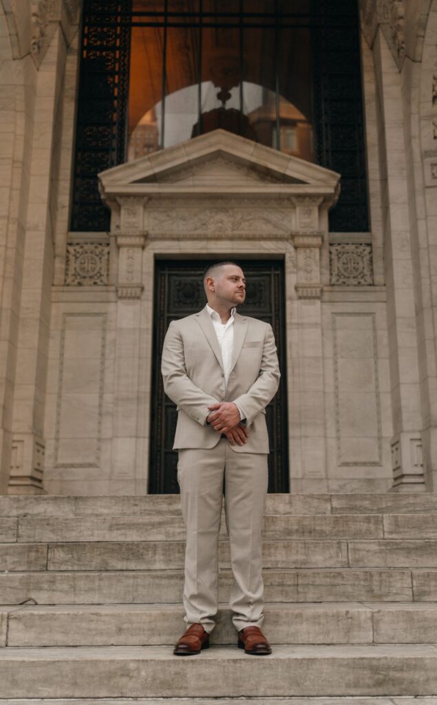 A groom dressed in a tan suit awaits his NYC city hall elopement on the front steps. 