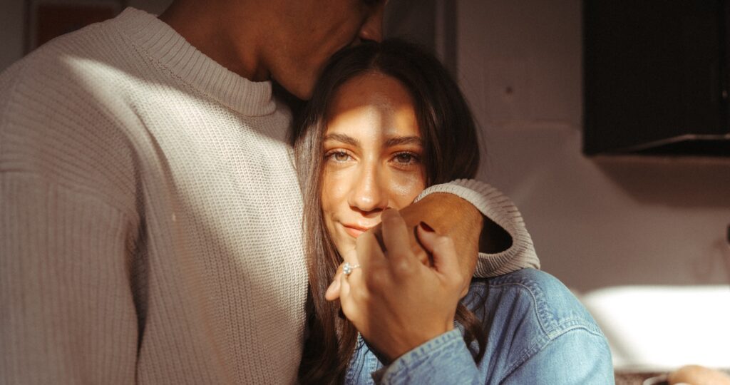 A young brunette woman leans into her fiance's chest as a sunbeam is cast across her face in an image taken by a NYC engagement photographer. 