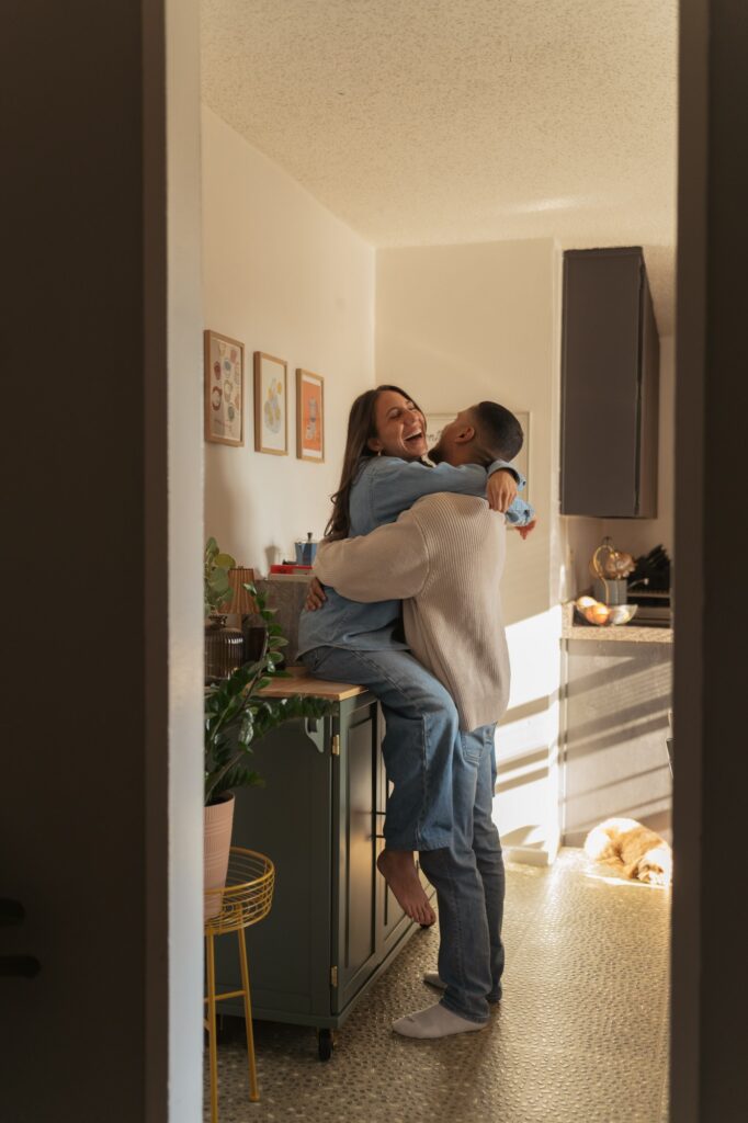 A couple laugh together & embrace on their kitchen counter during an in-home New York engagement session. 
