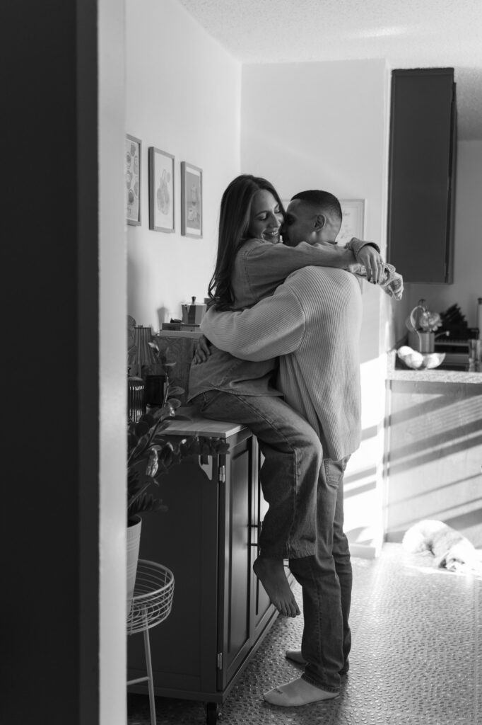 A black and white photo of a couple embracing on their kitchen counter during a New York engagement shoot in their Bronx apartment. 