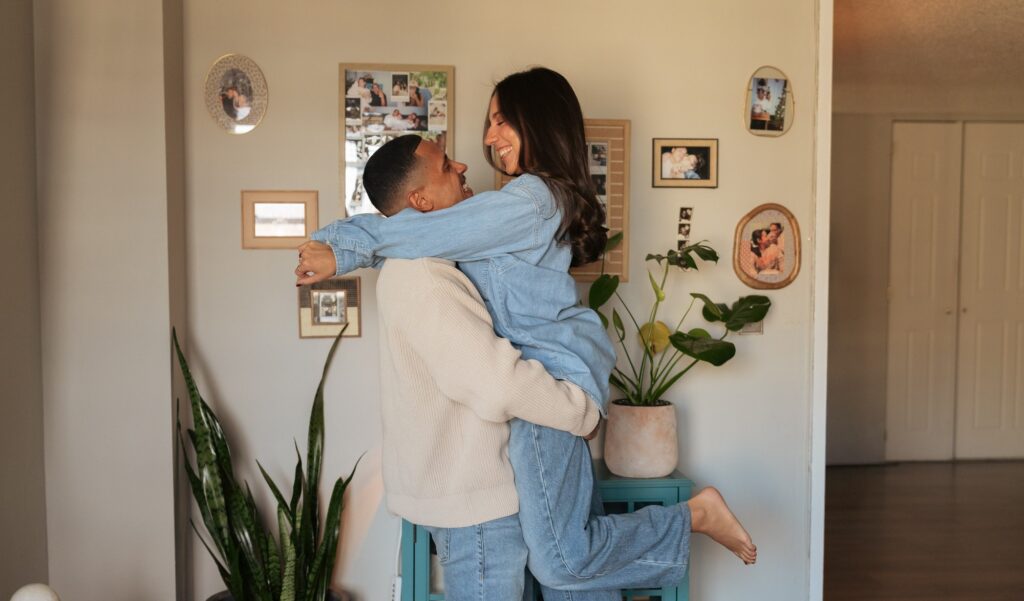 A man in a cream sweater lifts his fiancee in their living room during engagement pictures NYC.