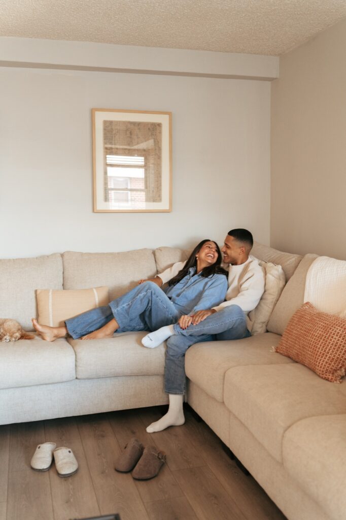 A couple lounges on their cream sofa in their Bronx apartment during in-home engagement pictures NYC. 